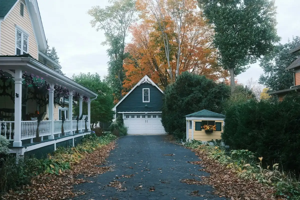 A long residential driveway running between two houses, lined with fallen autumn leaves and turning maples.