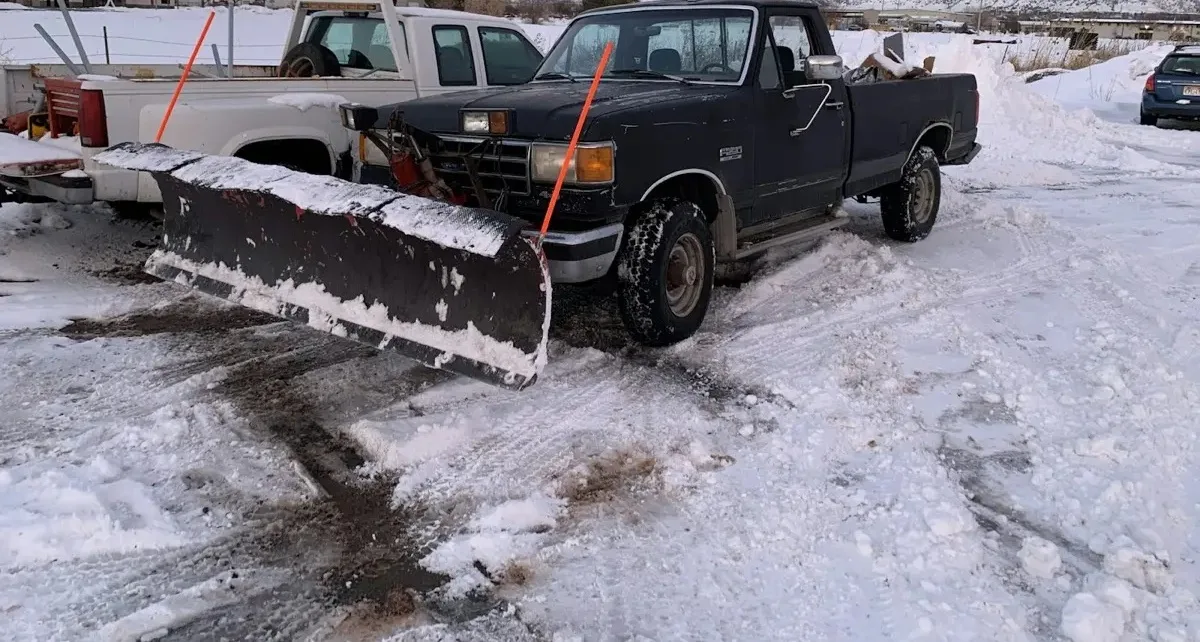 A pickup truck with a snow plow blade mounted on the front, standing on cleared snowy ground.